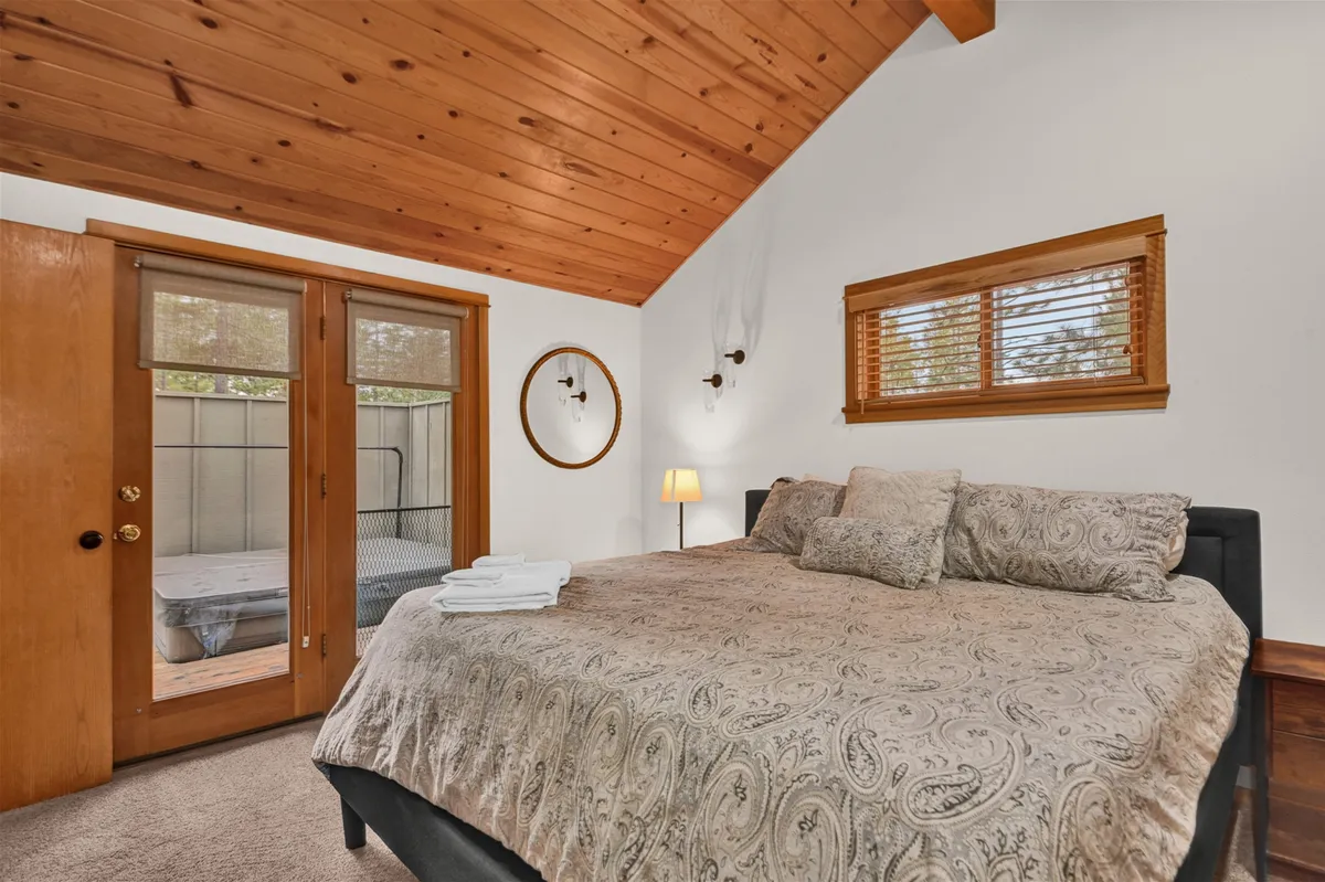 This image displays a primary bedroom with a king-sized bed adorned with decorative bedding, a wooden ceiling, sliding doors leading out to an outdoor area, a circular mirror, and a bedside table with a lamp. Natural light fills the room, and the sliding doors suggest direct access to the hot tub area outside.