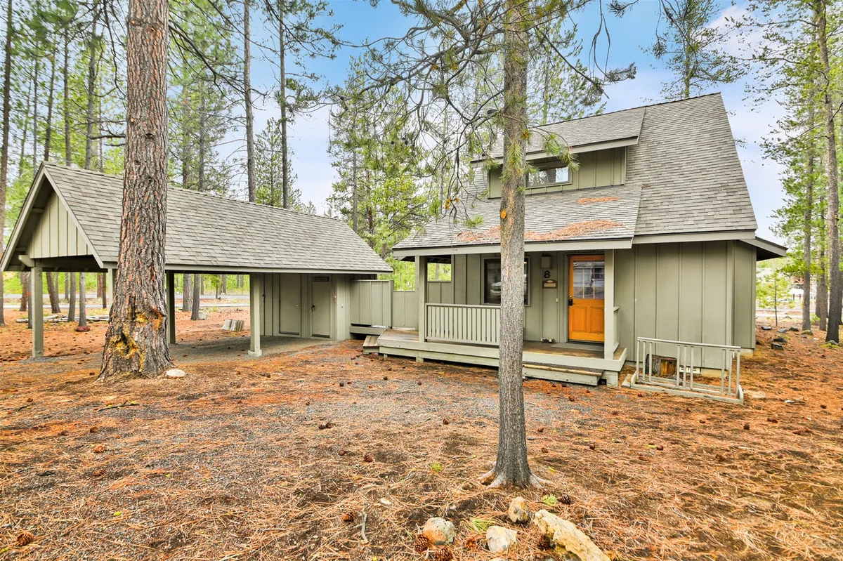 This image features the cabin's exterior nestled among tall pine trees, with a carport visible and the natural landscape surrounding the home. The setting conveys immediate access to the outdoors, perfect for adventure seekers.