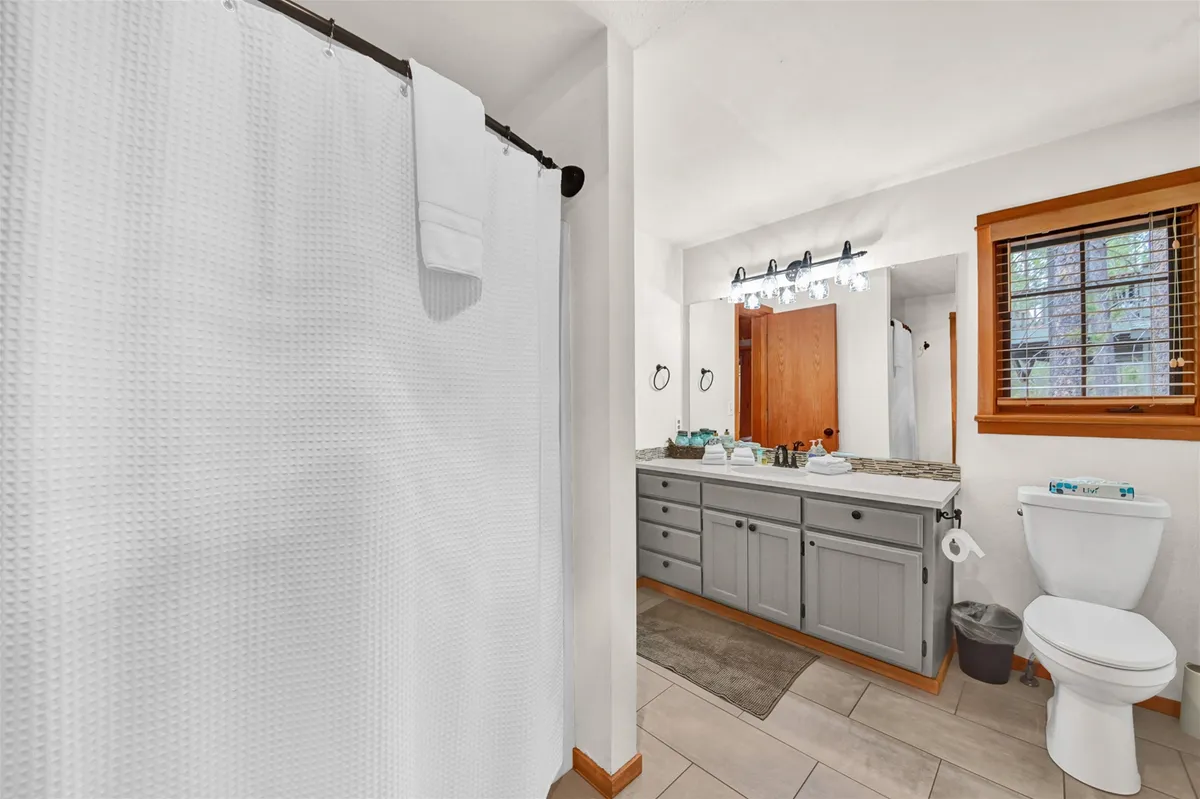 An inviting bathroom scene with a vanity and sink, a shower curtain suggesting a tub or shower, a toilet, and a window letting in natural light. The space is bright, clean, and practical—ideal for morning or evening routines.