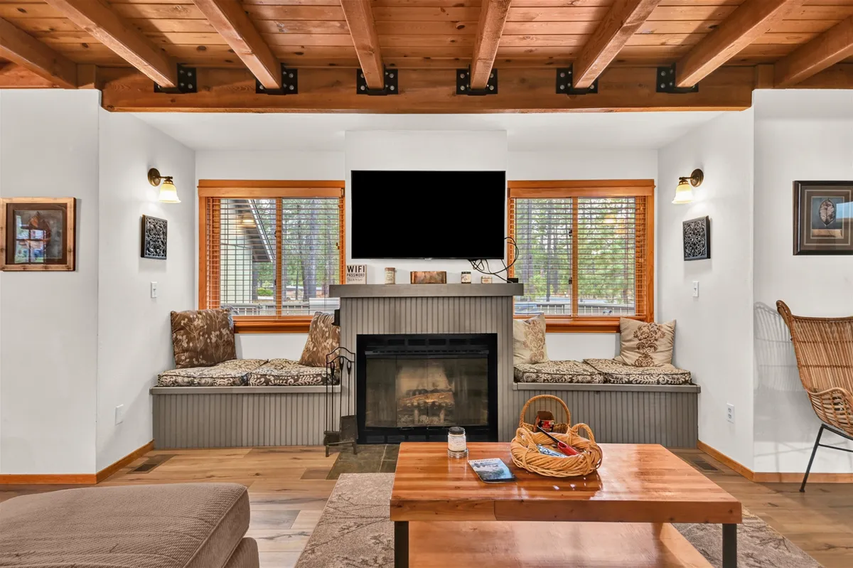 A cozy living room featuring a glowing wood fireplace, inviting window seating drenched in natural light, and a spacious coffee table perfect for gathering. The space feels updated, warm, and ideal for relaxing, reading, or spending quality time with loved ones.
