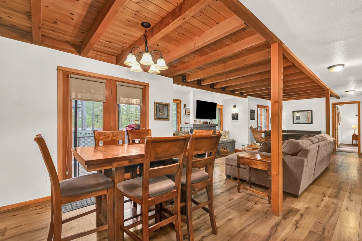 A multi-functional great room including a dining table, comfortable sofa, exposed wooden beams, a sliding door to the outside, and a prominently placed television. The room is ideal for shared meals, movie nights, and celebrations in air-conditioned comfort.