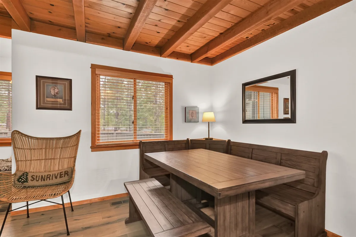 A spacious, welcoming dining area featuring a large wooden table surrounded by seating, with a woven 'SUNRIVER' chair adding a local touch. Large windows with blinds allow natural light to fill the space, and wooden beams on the ceiling evoke a rustic, communal feel. The overall mood is warm and inviting, perfect for shared meals and gatherings.