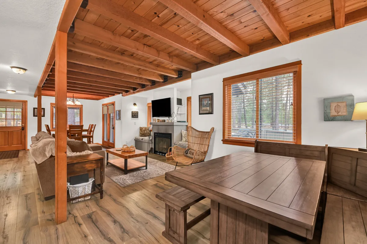 The image features a warm dining area with a wooden table, surrounding chairs, a glowing fireplace, exposed beams, and large windows that fill the room with natural light. A cozy rocking chair adds to the inviting atmosphere, and the scene is set for family meals or game nights.