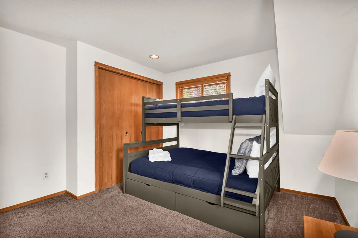 This image shows a cozy bedroom with a wooden bunk bed, a side lamp, and a wooden closet door. The room is simply decorated and designed for practicality, providing comfortable sleeping arrangements for kids or groups.