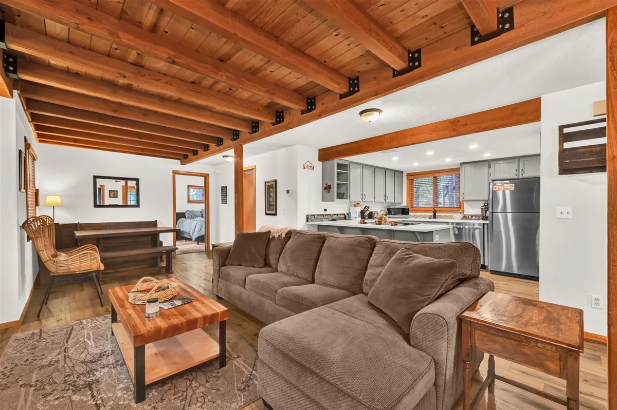 This image showcases a cozy open-concept living area featuring a plush sofa, visible wooden beams, a kitchen area with stainless steel appliances, and a coffee table. The dining table is set nearby, and there is a welcoming interplay of spaces, emphasizing the seamless flow from living to dining to kitchen.